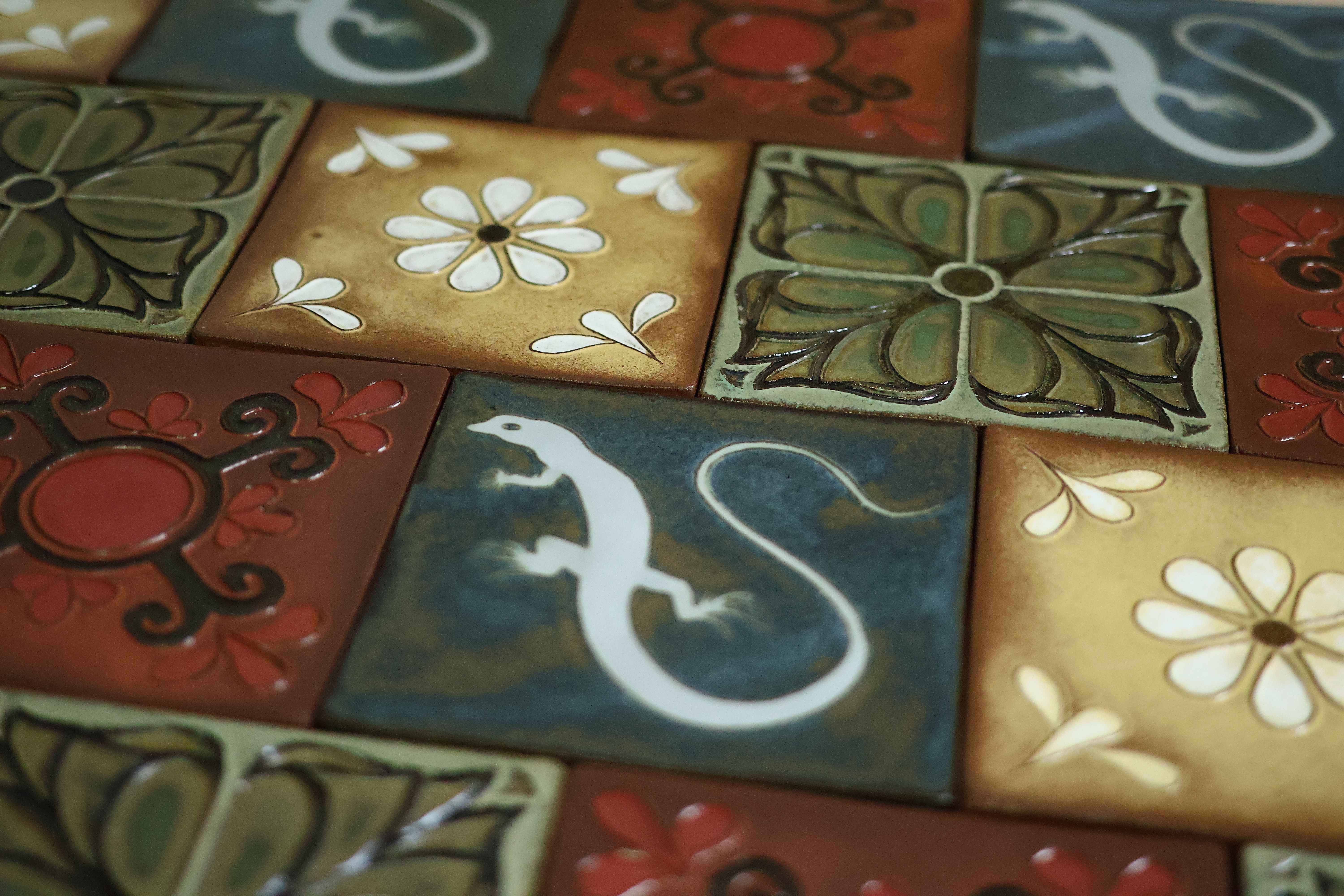 Angled close-up of glossy Courtyard tiles — white daisies on golden grounds, green embossed florals, rust-red medallions, and a deep-blue tile with a white lizard