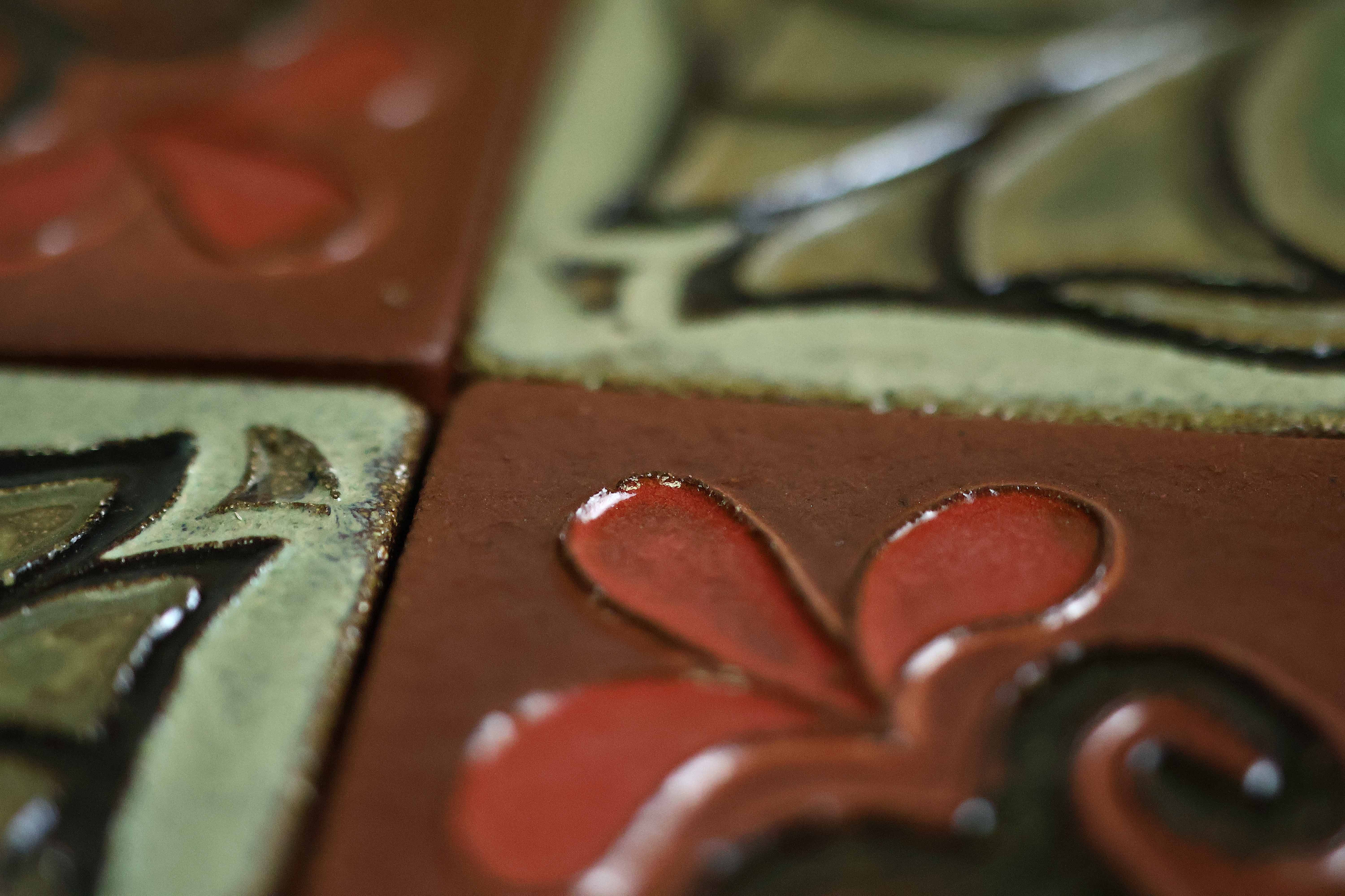 Macro close-up of a terracotta Courtyard tile with a raised red petal motif and black scrollwork beside a green scrollwork tile